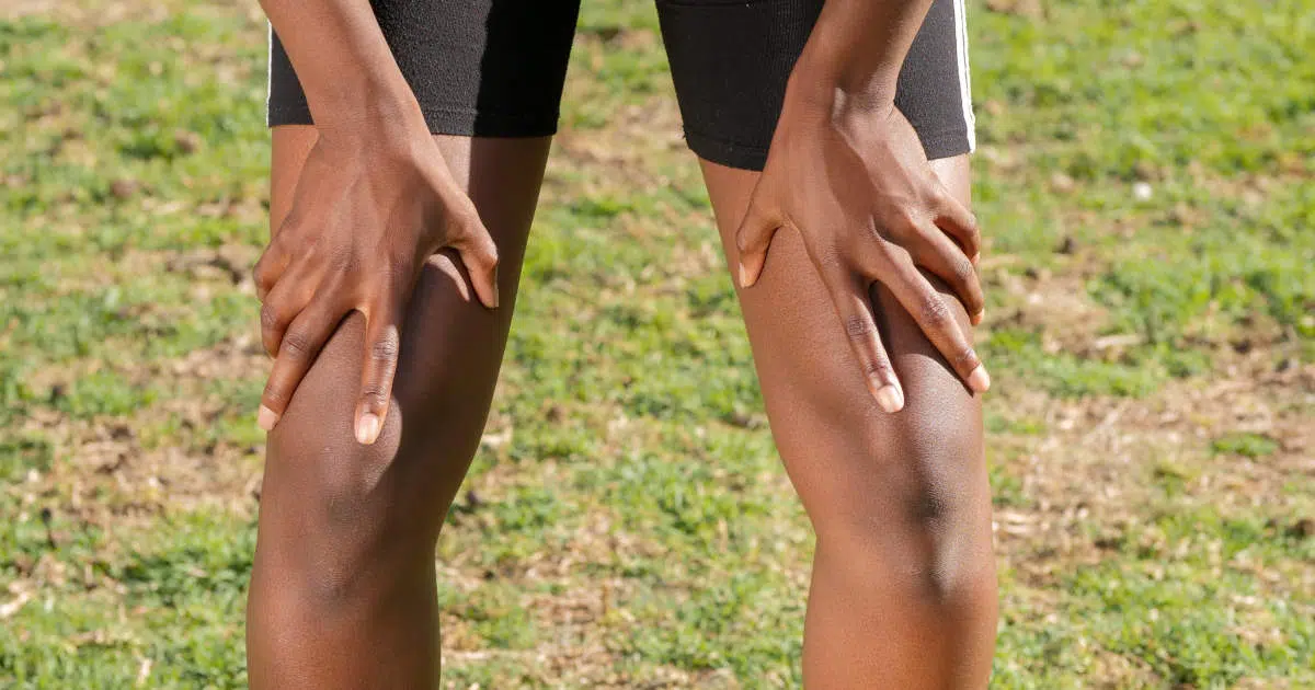 Eine Person steht mit kurzen, schwarzen Hosen auf einer Wiese. Sie hat die Hände auf beide Knie abgelegt und beugt sich vorn über.