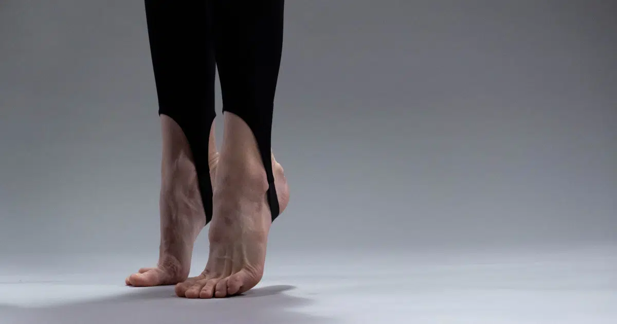 Zwei Füße stehen auf den Fußballen. Die Füße sind nackt, aber die Person trägt eine schwarze Turnhose, die jeweils mit einer Schlaufe um die Ferse festgemacht ist.