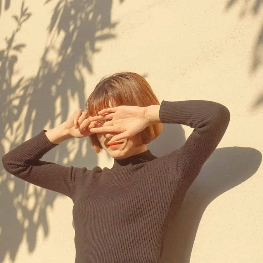 Eine junge Frau steht in der Sonne und hält sich die Hände vor Augen. Hinter ihr fallen Schatten von Bäumen.