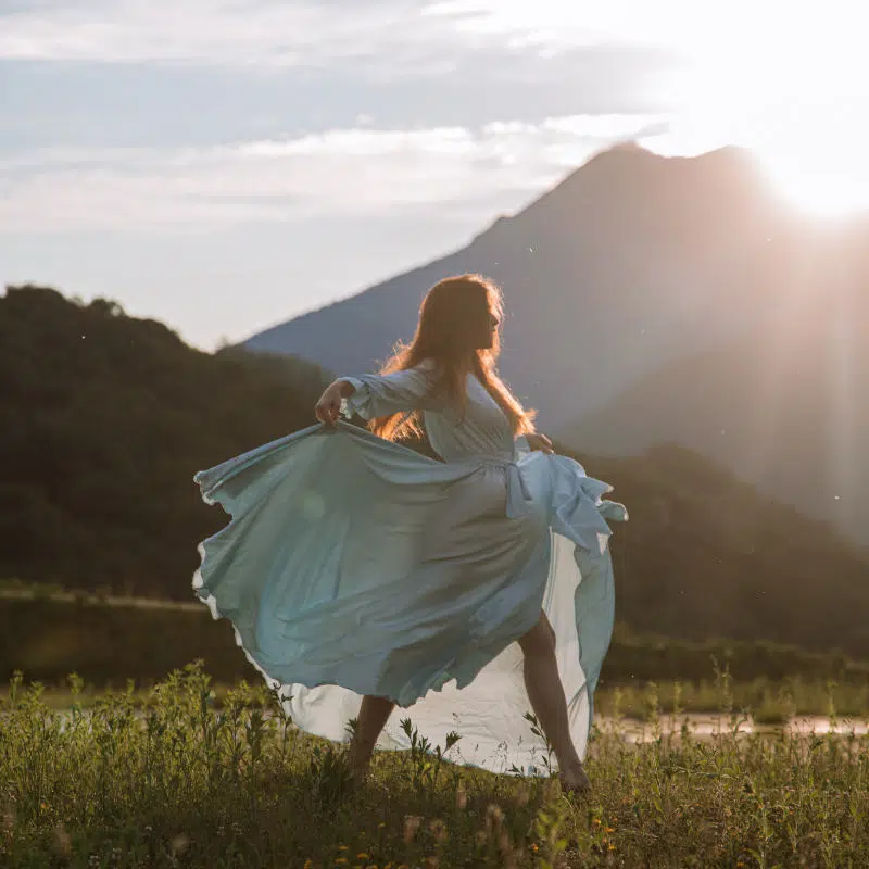 Eine junge Frau läuft im luftigen Kleid in der Sonne über eine grüne Wiese
