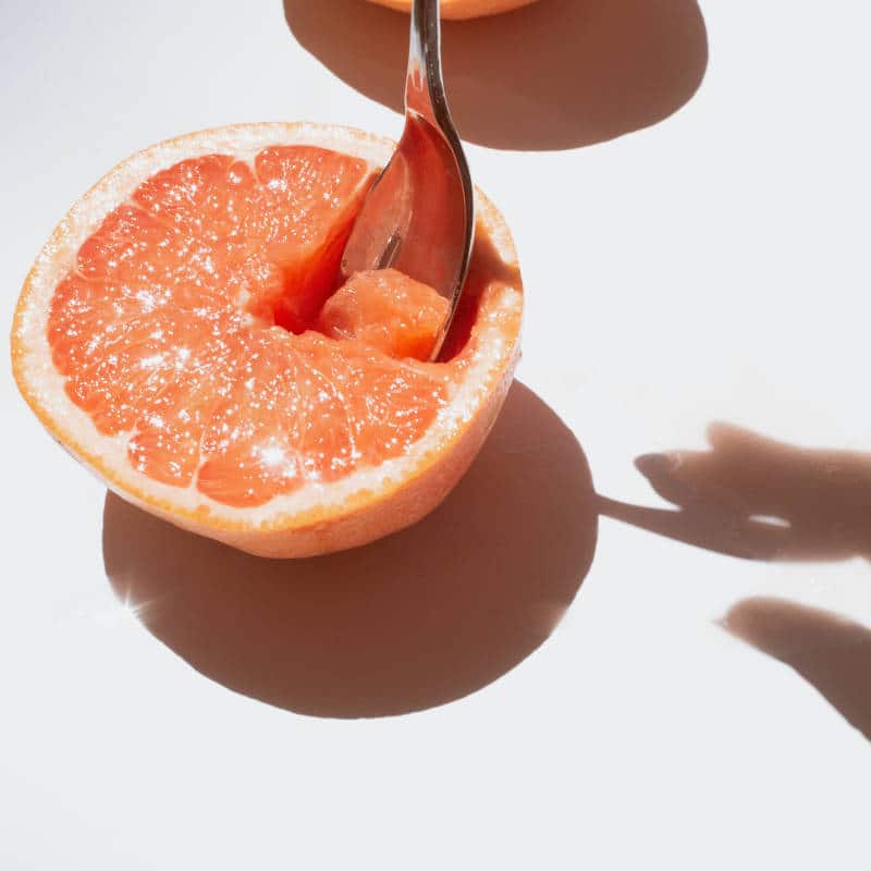 Eine aufgeschnittene Grapefruit liegt auf einem weißen Untergrund. Der Schatten einer Hand, die die Grapefruit löffelt, ist zu sehen.
