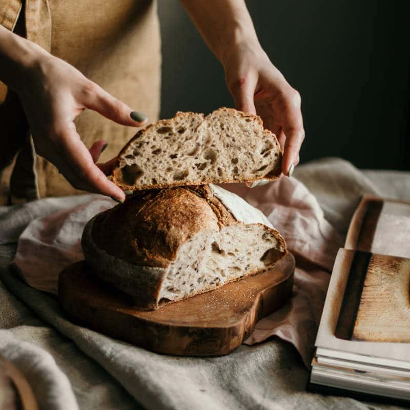 Eine Frau mit Schürze hält ihr fertiges Brot in den Händen. Es ist in zwei Hälften geteilt. Die eine Hälfte liegt auf einem Brotbrett.