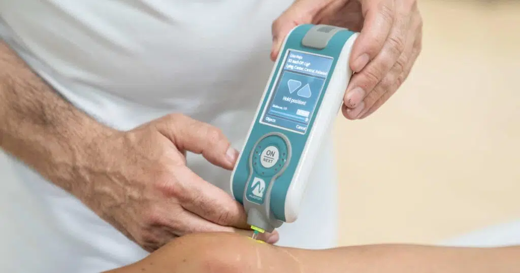 Mit einem Myometer, dass von zwei Händen gehalten wird, wird die Muskelspannung an einem Knie gemessen, dass unten im Bild zu sehen ist.