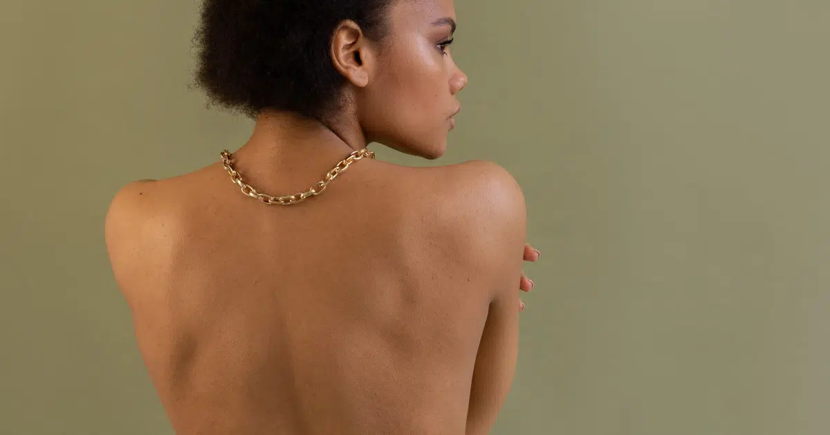 Ausschnitt vom Schulterbereich einer Person. Sie ist von hinten zu sehen und blickt nach rechts, sodass ihr Profil sichtbar ist. Ihr Oberkörper ist nackt, sie trägt eine goldene Halskette. Ihre Haare sind schwarz.
