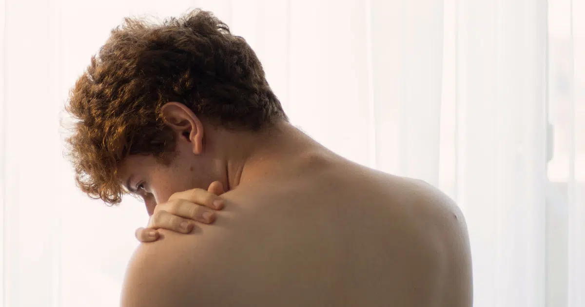 Ein Jugendlicher steht mit freiem Oberkörper vor einem Fenster und hat seinen Rücken dem Betrachter zugewandt. Sein Kopf ist geneigt und man kann einen leichten Buckel oder Rundrücken erkennen.