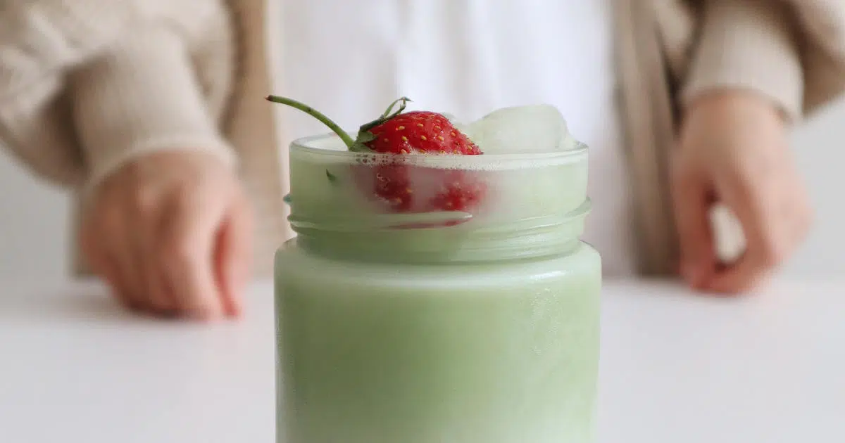 Ein Matcha-Mandelmilch-Latte ist mit Eiswürfeln und einer Erdbeere im Glas abgebildet. Dahinter steht eine Frau. Von ihr sieht man nur die Hände auf der Tischplatte und ihr Oberteil.