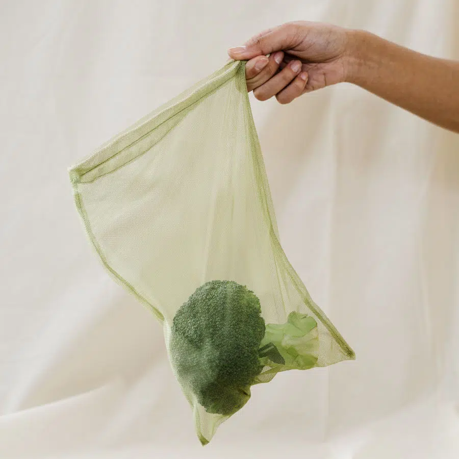 Eine Frauenhand hält ein Gemüsenetz mit einem Brokkolikopf vor ein weißes Tuch.