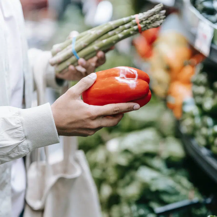 Eine Frau hält in der linken Hand einen Bund grünen Spargel und in ihrer rechten eine rote Paprika. Sie steht an einem Gemüsestand auf dem Markt