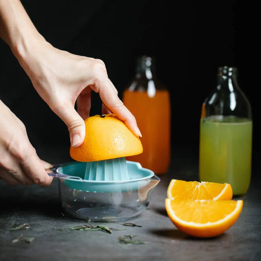 Eine Frauenhand presst Orangenhälften für Frühstückssäfte mit einer Zitruspresse aus. Im Hintergrund stehen zwei Flaschen mit fertigen Säften.