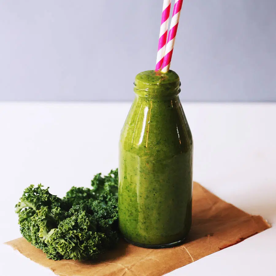 Ein grüner Smoothie mit Grünkohl ist in einer Glasflasche auf einem Tisch. Aus der Glasflasche ragen zwei pink-weiße Strohalme