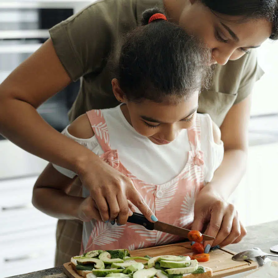 Eine Mutter steht mit ihrer Tochter in der Küche und schneidet Avocado und Tomaten für ein Sandwich.