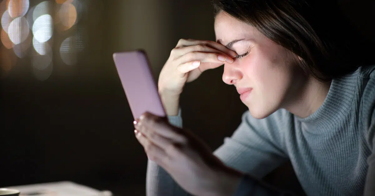 Eine Frau hält im Dunkeln ihr Smartphone hoch. Das weiße Licht des Displays erhellt ihr Gesicht. Sie kneift ihre Augen zu und mit den Fingern der anderen Hand greift sie an die Innenwinkel ihrer Augen.