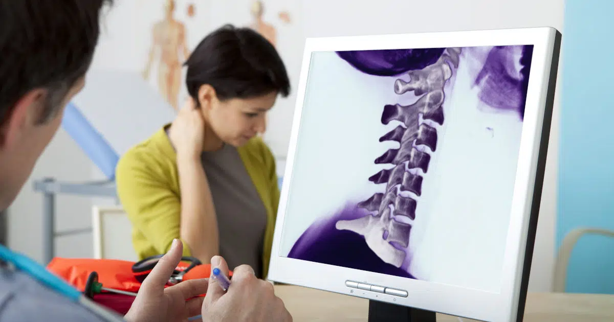 Eine Person sitzt beim Arzt. Sie hält sich mit der rechten Hand den Nacken fest. Der Arzt ist im linken Bildausschnitt kaum zu sehen. Vor ihm steht ein Bildschirm mit dem Röntgenbild einer Halswirbelsäule.