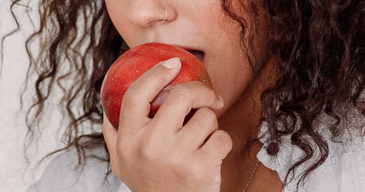 Eine Frau hält einen großen roten Apfel in ihrer Hand. Sie öffnet Kiefer und Mund, um einen großen Bissen mit ihren Zähnen zu nehmen.