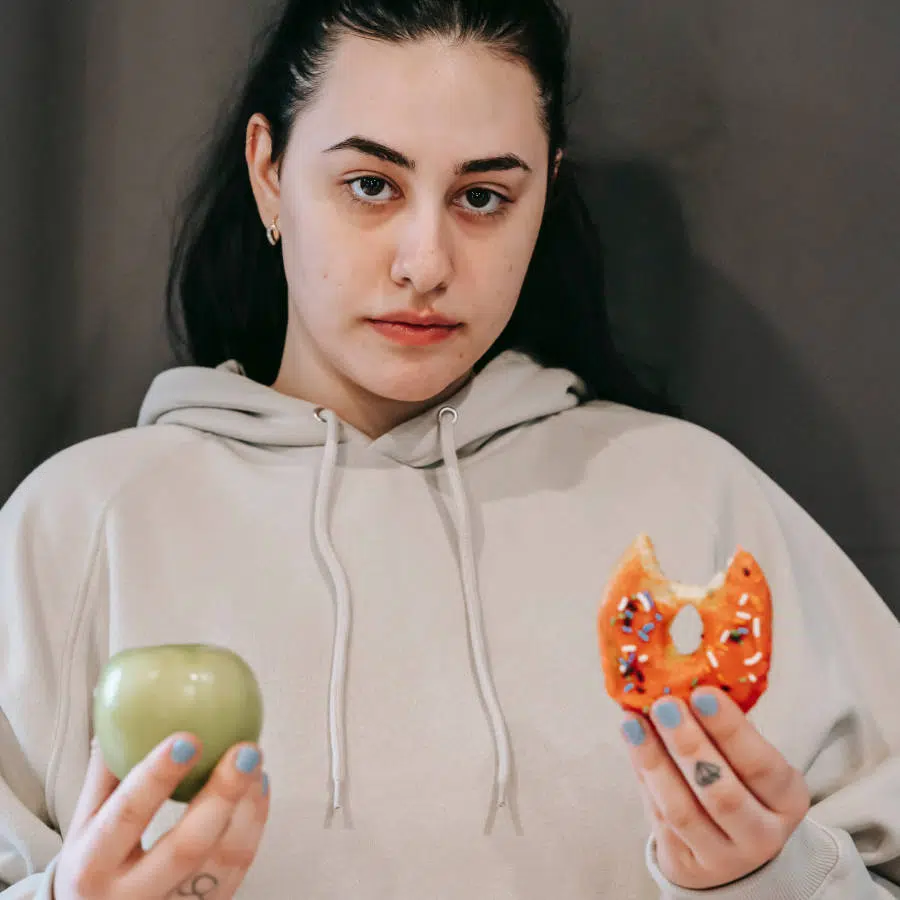 Eine leicht übergewichtige Frau hält in der linken Hand einen angegessenen Donut und in der rechten Hand einen grünen Apfel.