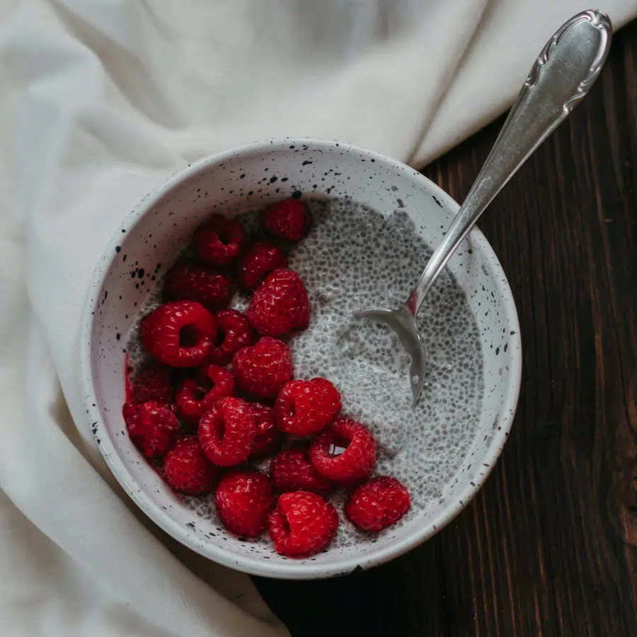 Eine Tonschale mit Chiapudding und frischen Himbeeren steht auf einem Tisch. Ein Löffel steckt im Pudding und um die Schale ist eine weiße Tischdecke gelegt.