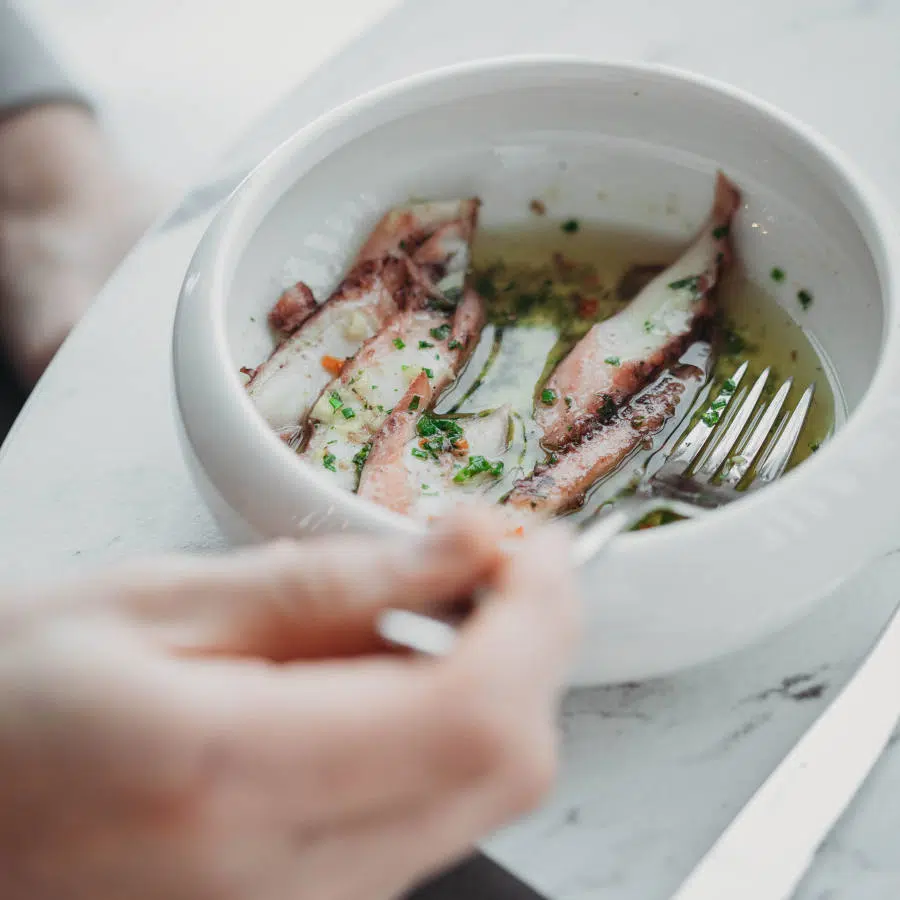 Ein Fischgericht in einer Schüssel steht auf einem weißen Tisch. Eine Person hält mit der rechten Hand eine Gabel in der Hand und isst.