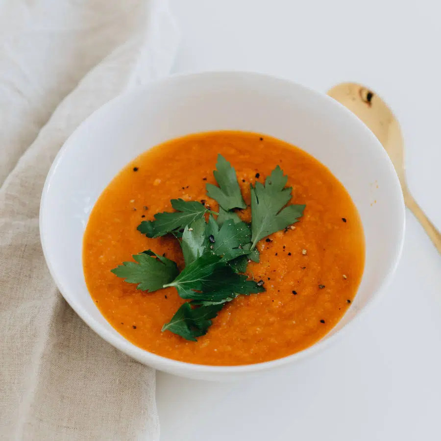 Eine orangene Kürbissuppe in einer weißen Schale steht auf einem weißen Tisch. Sie ist mit frischer Petersilie garniert.