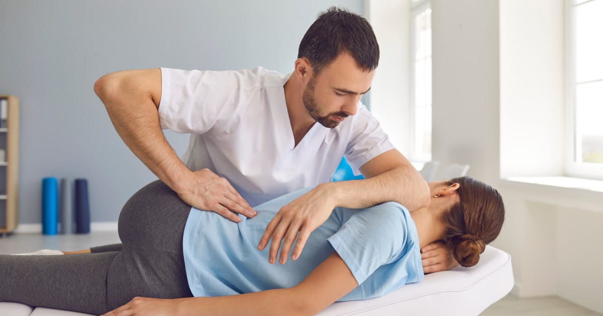 Ein Therapeut führt eine Mobilisierung der Manuellen Therapie an einer Frau aus. Der Therapeut trägt ein weißes T-Shirt und übt mit der rechten Hand Druck auf den Hüftbereit der Frau aus. Die Frau im hellblauen T-Shirt liegt seitlich auf einer Behandlungsliege und hat das linke Bein angewinkelt.