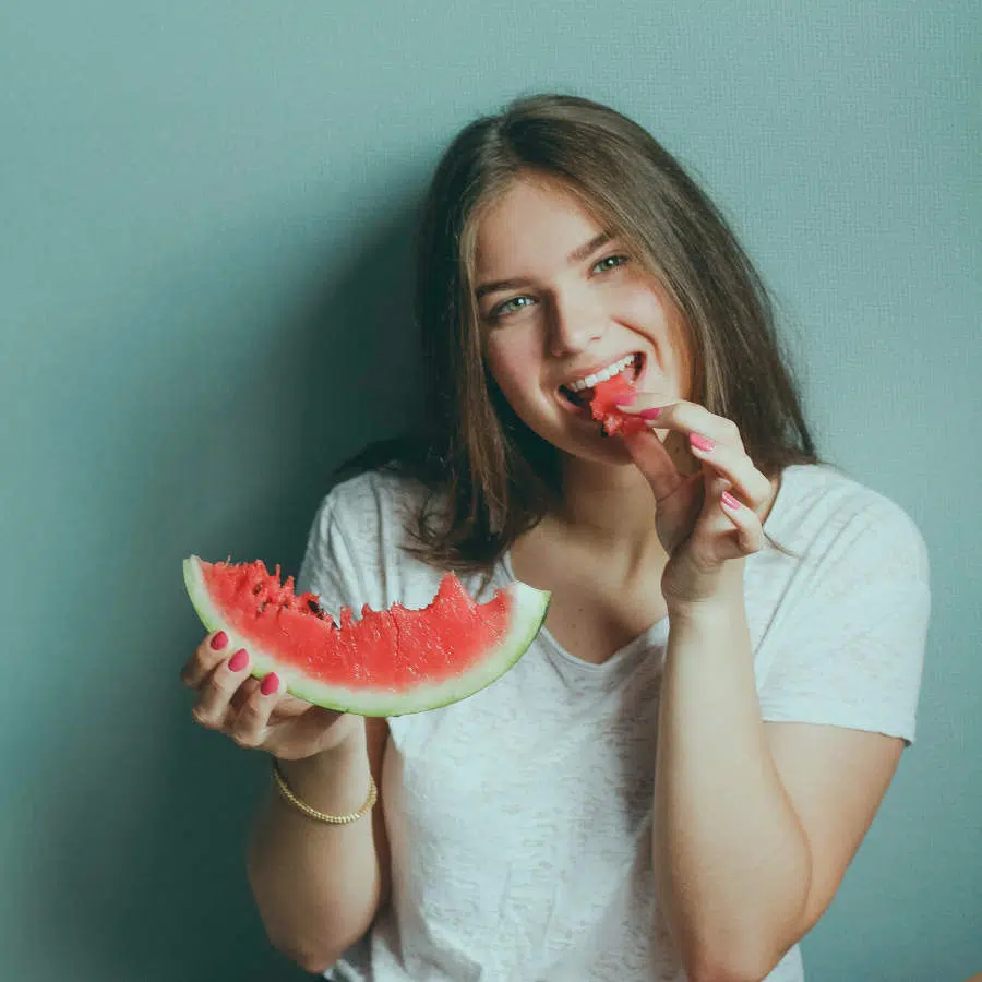 Eine junge Frau hält in der rechten Hand ein großes Stück Wassermelone und beißt in ein kleines Stück rein.