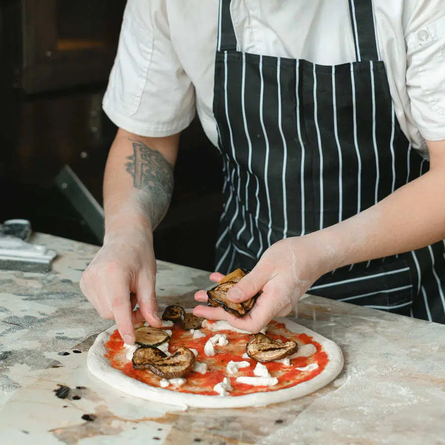 Ein Koch mit Schürze belegt eine Pizza mit rein pflanzlichen Zutaten.