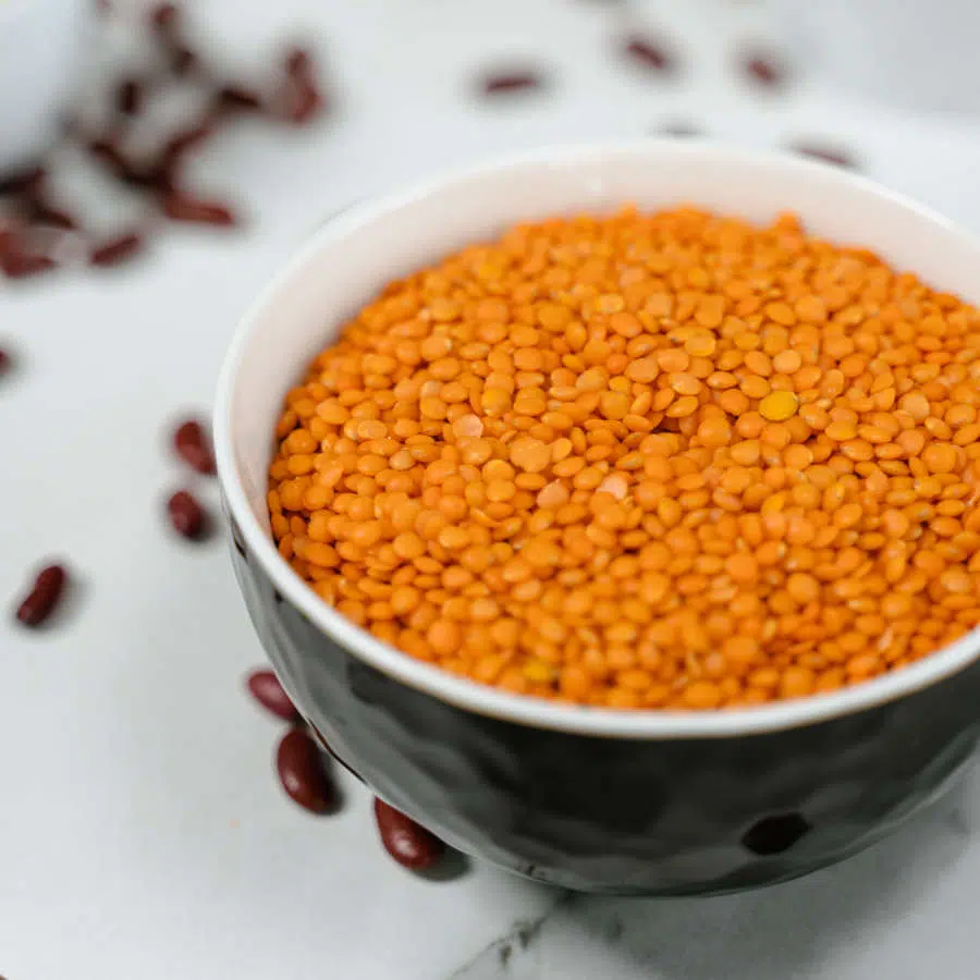 Rote Linsen sind in einer schwarzen Schale. Drum herum liegen rote Bohnen auf einem Tisch verteilt.