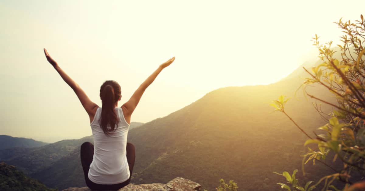 Man sieht eine Frau mit einem weißen Topvon hinten, sitzend beide Arme über den Kopf gestreckt. Sie befindet sich draußen in der Natur in einer hügeligen Landschaft. Vor ihr versteckt sich die Sonne hinter einem Berg.