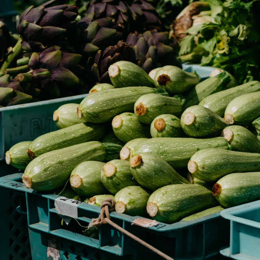 Viele hellgrüne Zucchini liegen an einem Marktstand übereinander gestapelt. Dahinter sind viele Artischocken zu sehen.