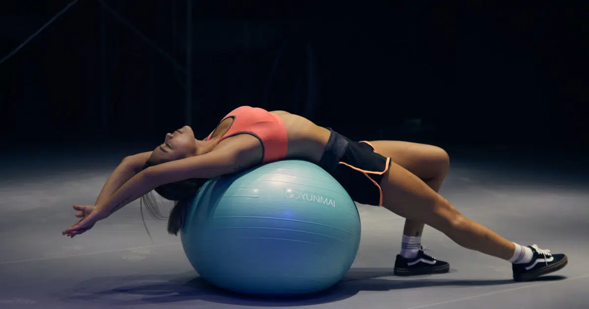 Eine Person liegt mit dem Rücken auf einem Gymnastikball. Sie hat die Arme nach hinten gestreckt und lässt sich passiv hängen. Dabei wird die Brustmuskulatur gedehnt. Die Beine sind nach vorne ausgestreckt, das linke Bein ist leicht angewinkelt, das rechte ist gestreckt.