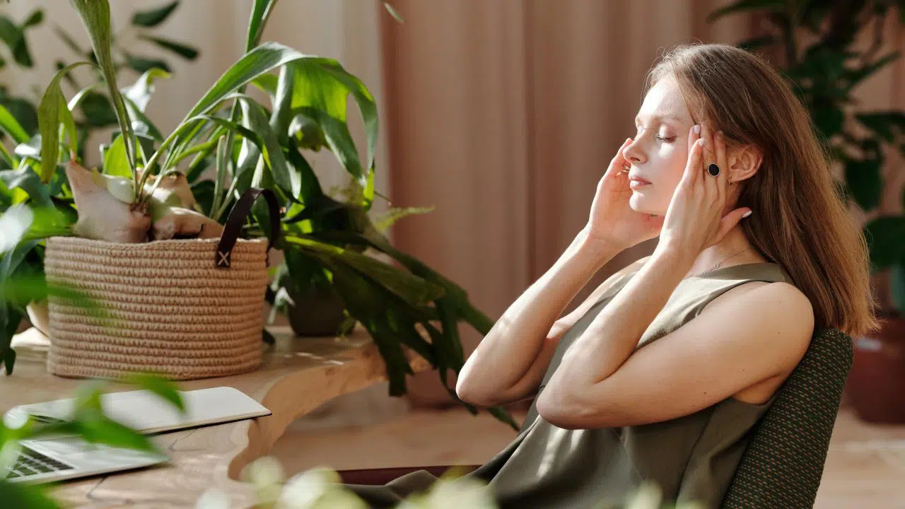 Eine Frau sitzt am Schreibtisch und massiert sich die Schläfen. Ihre Augen hälts sie dabei geschlossen.