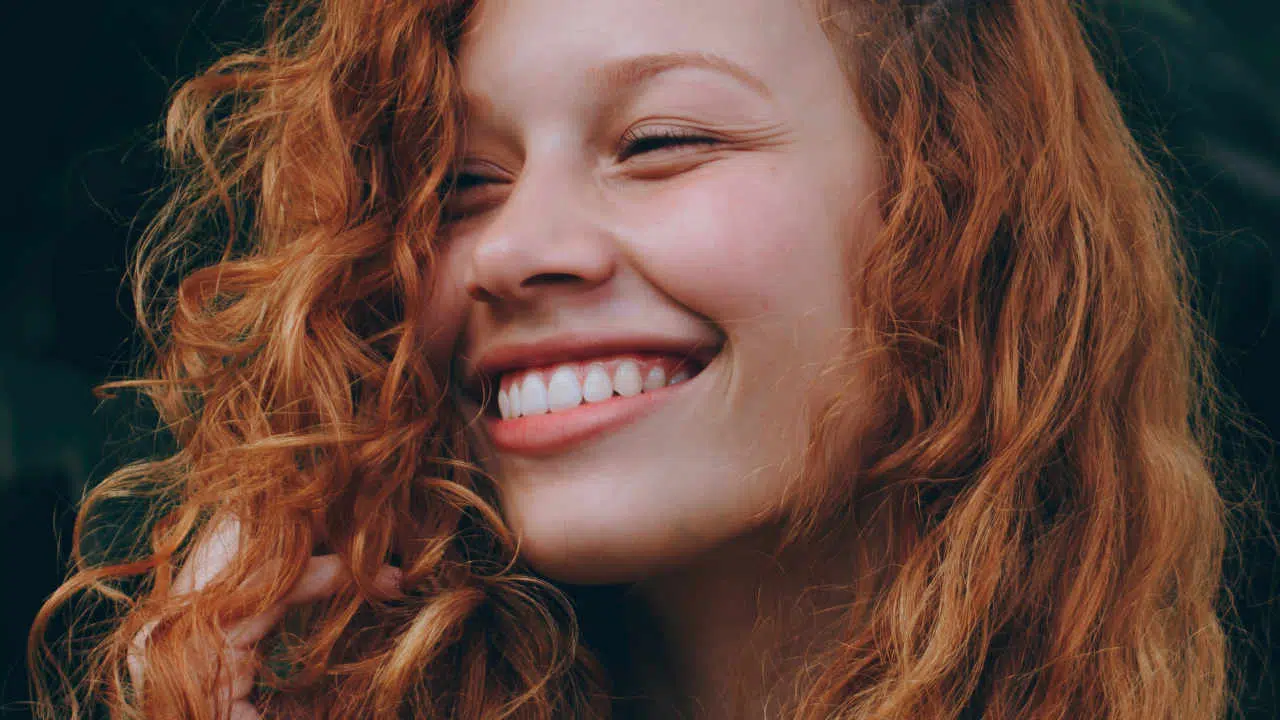 Eine rothaarige Frau hat ihren Kopf leicht nach rechts gedreht und strahlt über ihr ganzes Gesicht. Ihr Lächeln zeigt weiße, gepflegte Zähne.