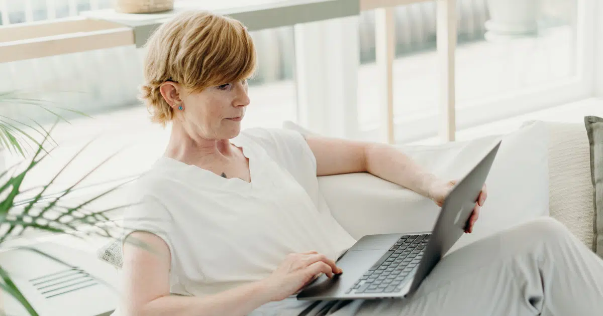 Das Foto zeigt eine Frau im weißen Shirt und mit rötlichen kurzen Haaren, die auf einer Couch vor einem aufgeklappten Laptop sitzt und die Beine übereinander schlägt.