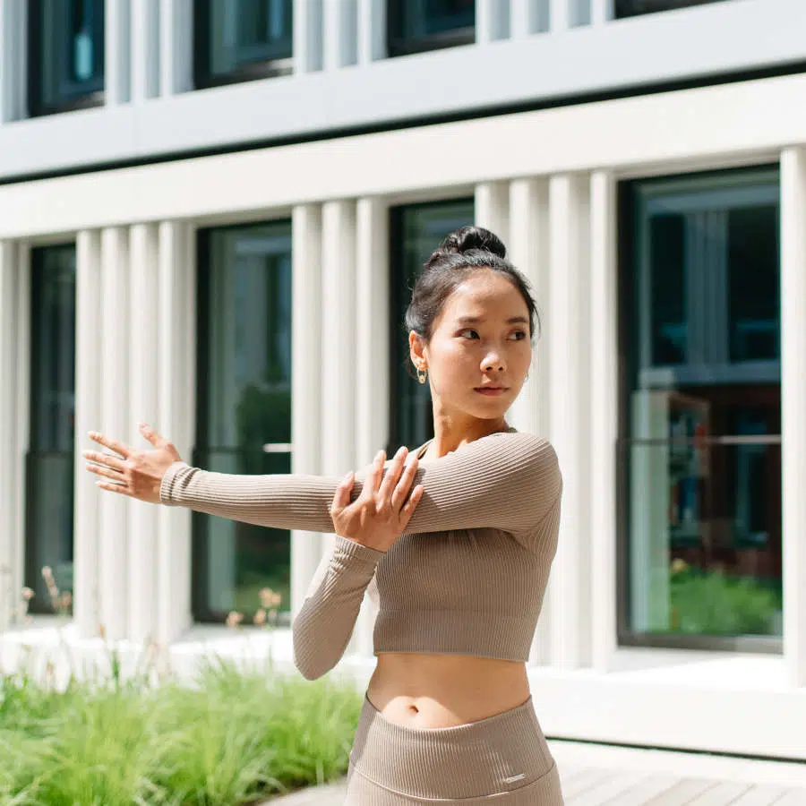 Eine junge Frau steht vor einem Gebäude und dehnt ihren linken Arm, indem sie ihn mit dem rechten Arm ausgestreckt nach rechts zieht.