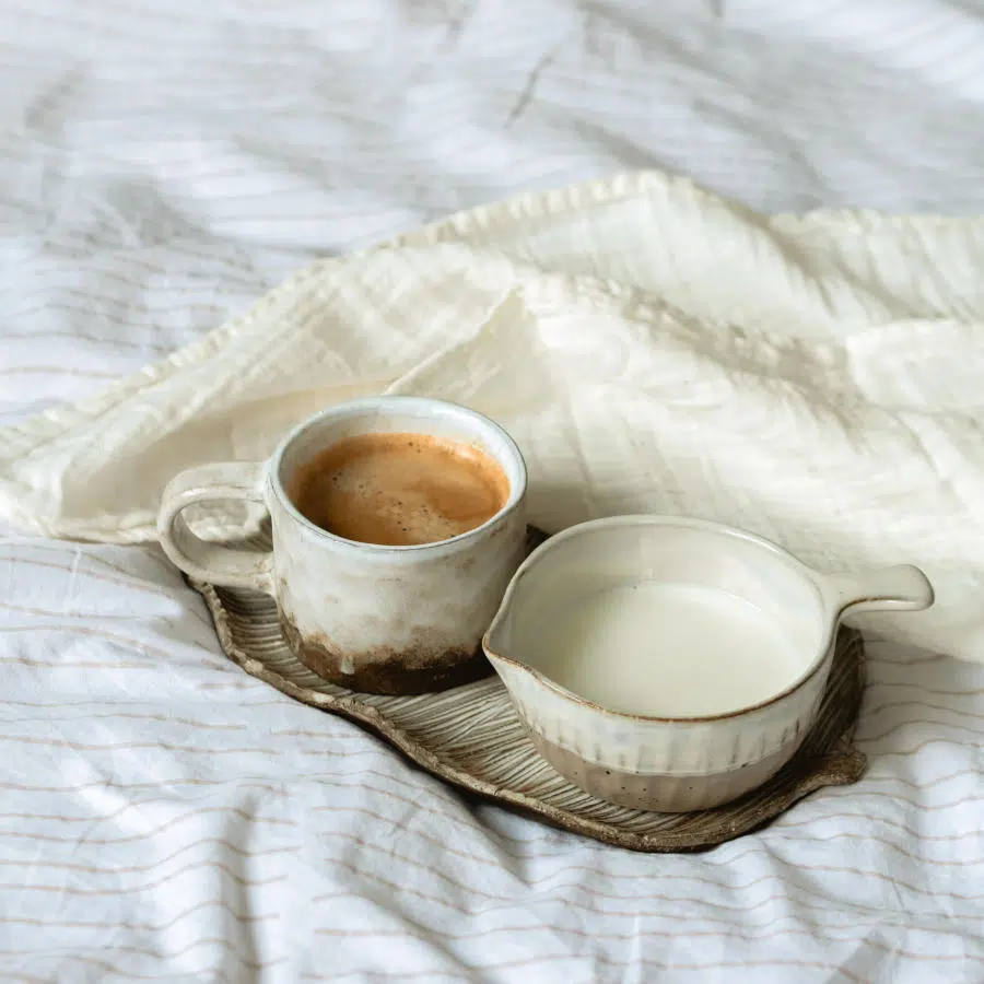 Eine Tasse Kaffee Creme steht zusammen mit einem Gefäß voll mit Pflanzenmilch nebeneinander auf einem weißen Bettbezug.