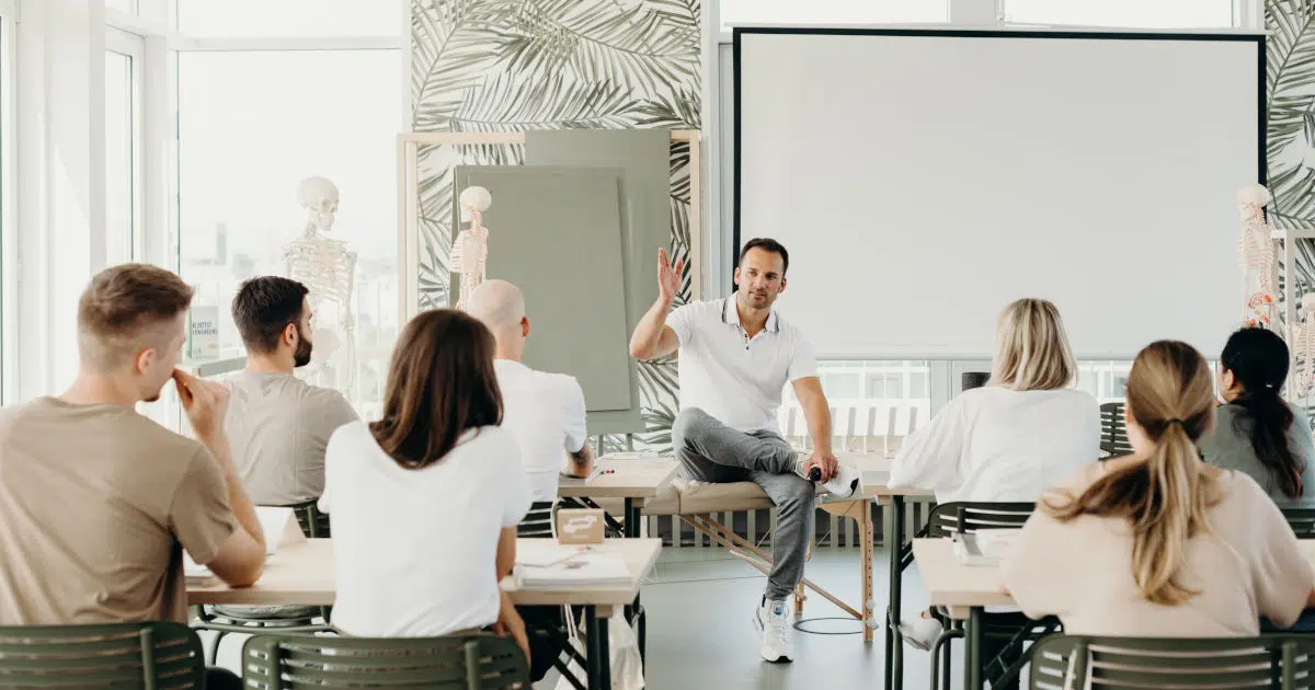 Ein Mann im weißen Poloshirt und grauer Sporthose sitzt mit überschlagenen Beinen vor einer Gruppe Ausbildungsteilnehmern. Im Hintergrund ist ein Whiteboard und ein Schulskelett zu sehen.