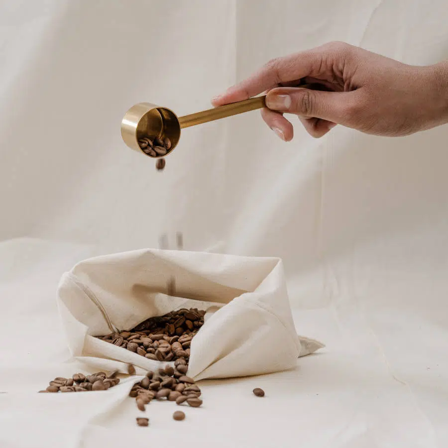 Ein weißer Leinensack mit gerösteten Kaffeebohnen steht auf einem Boden. Eine Hand entleert einen goldenen Löffel mit Kaffeebohnen über dem Leinensack