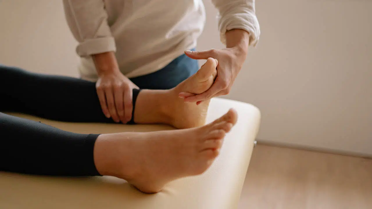 Auf einer Massageliege sind zwei nackte Füße. Dahinter steht ein Therapeut, der mit seinen Händen die Großzehe und den Vorfuß untersucht.