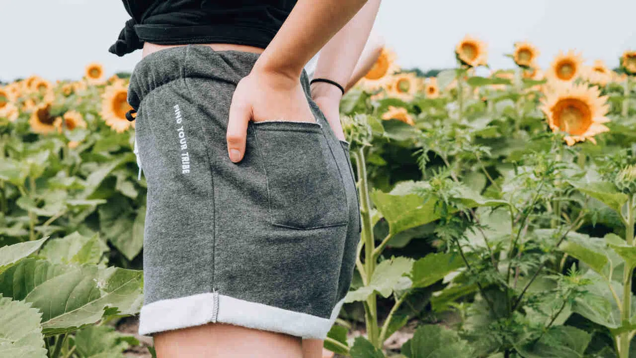 Eine Person steht vor einem Feld mit Sonnenblumen. Sie trägt eine kurze Hose, die am Oberschenkel endet, und sie hat ihre Hände in die Taschen über dem Gesäß gesteckt.