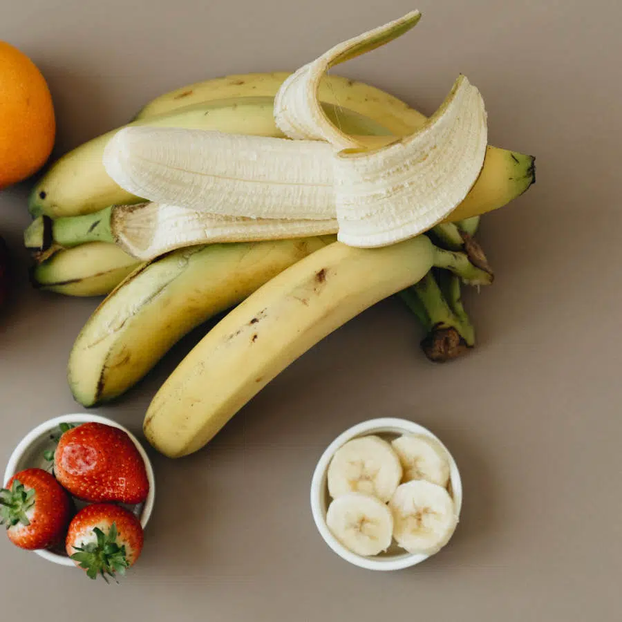 In der Mitte des Bildes liegen mehrere Bananen aufeinander, von denen die oberste halb geschält ist. Darunter stehen zwei kleine Schüsseln. In der linken sind drei Erdbeeren und in der rechten vier Scheiben Bananen enthalten.