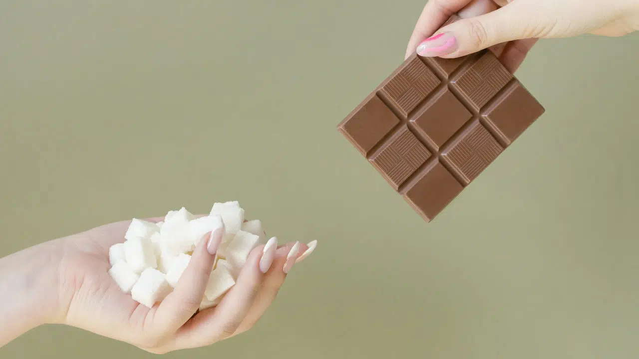 Eine Hand rechts oben im Bild hält eine Tafel Schokolade ins Bild, während eine Hand von links unten viele weiße Würfelzucker ins Bild hält.