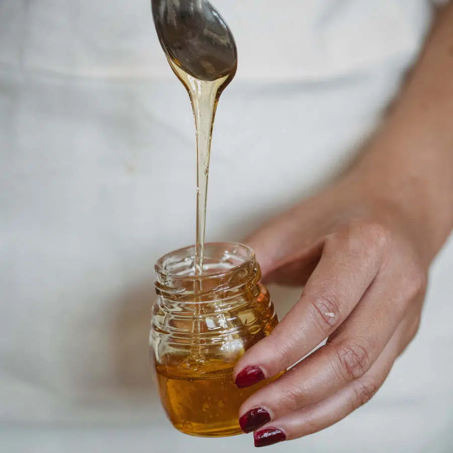 Die linke Hand einer Frau hält ein kleines Glas mit Sirup, während ihre rechte Hand Sirup von einem Löffel ins Glas laufen lässt.