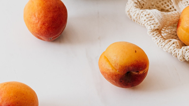 Bildausschnitt eines Tisches mit weißer Tischdecke, auf dem ein Teil eines Einkaufsnetzes zu sehen ist. Aus diesem sind ein paar gelbe Aprikose rausgerollt und liegen verstreut auf der Decke.