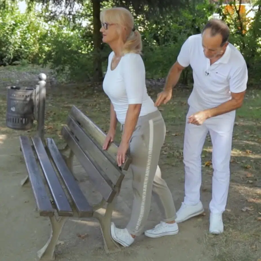 Roland Liebscher-Bracht steht neben einer Frau in Trainingskleidung und erklärt ihr die erste Übung an der Parkbank.