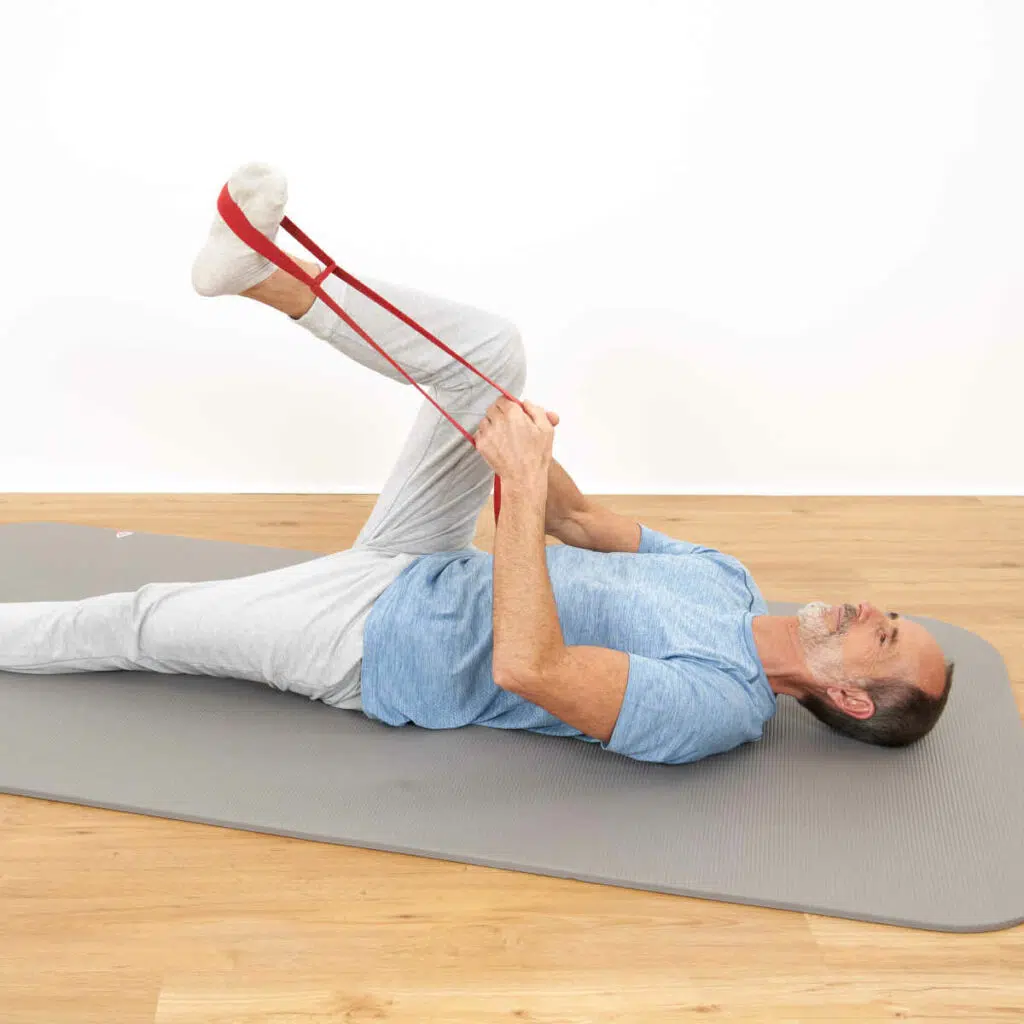 Roland Liebscher-Bracht liegt mit dem Rücken auf einer grauen Trainingsmatte. Ein Bein hat er ausgestreckt, das andere Bein angehoben und angewinkelt. Um seinen Fuß ist eine rote Schlaufe gewickelt, mit der er den Fuß zu sich heranzieht.