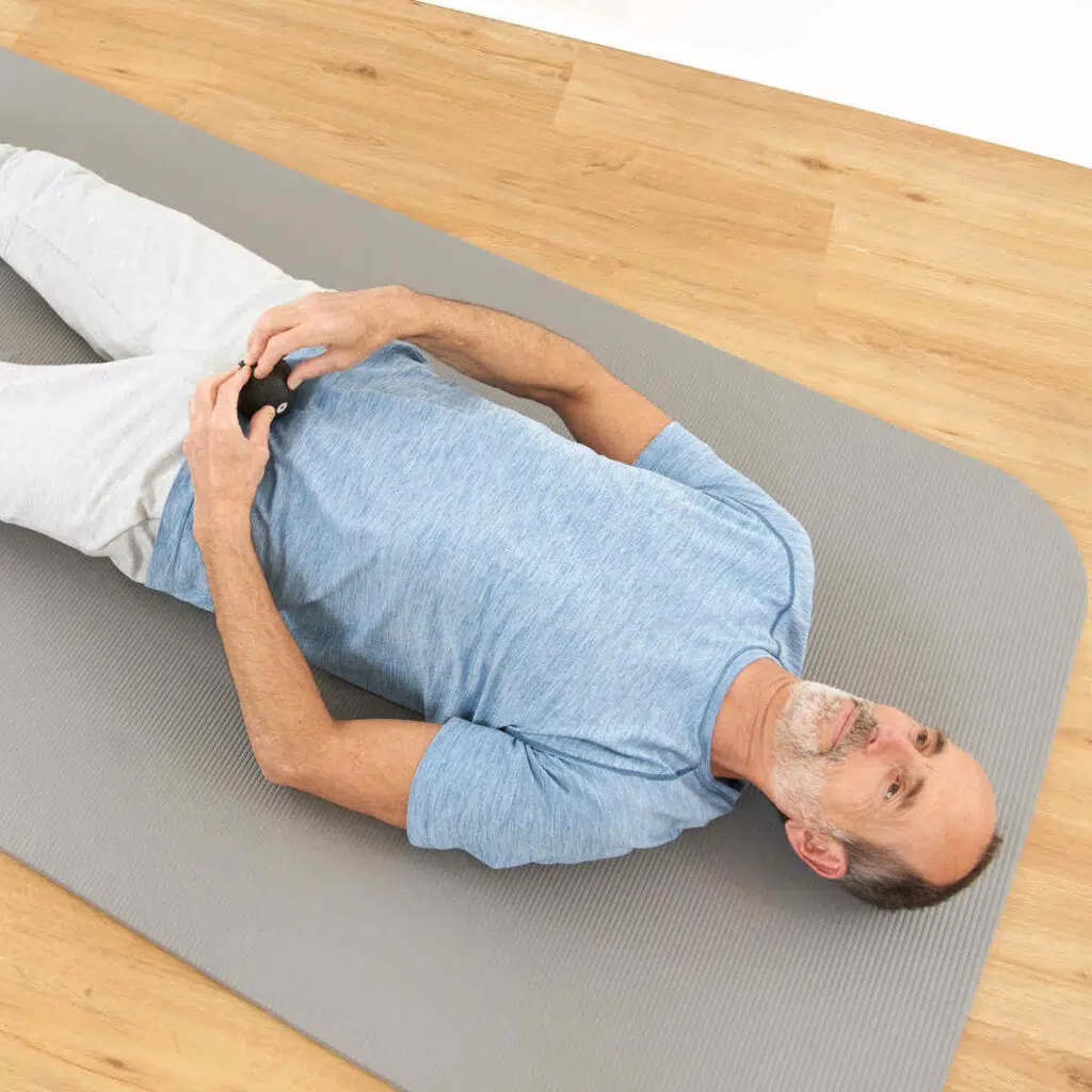 Auf einer grauen Trainingsmatte liegt Roland Liebscher-Bracht. Mit seinen Händen hält er eine kleine Faszienkugel an sein Schambein.