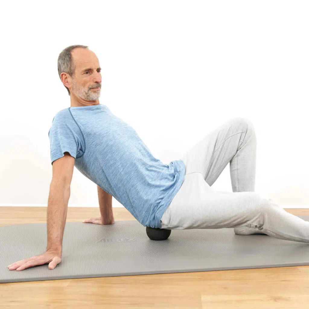 Roland Liebscher-Bracht sitzt mit dem Gesäß auf einer mittelgroßen Faszienkugel. Ein Bein hat er ausgestreckt, das andere Bein ist angewinkelt und er stützt sich nach hinten mit seinen Armen ab.