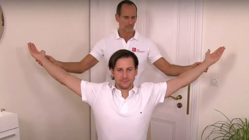 Ein Mann sitzt auf einem Stuhl und hat seine Arme seitlich ausgestreckt. Hinter ihm steht Schmerzspezialist Roland Liebscher-Bracht und bringt die Arme des Manns leicht nach hinten in eine Dehnung.