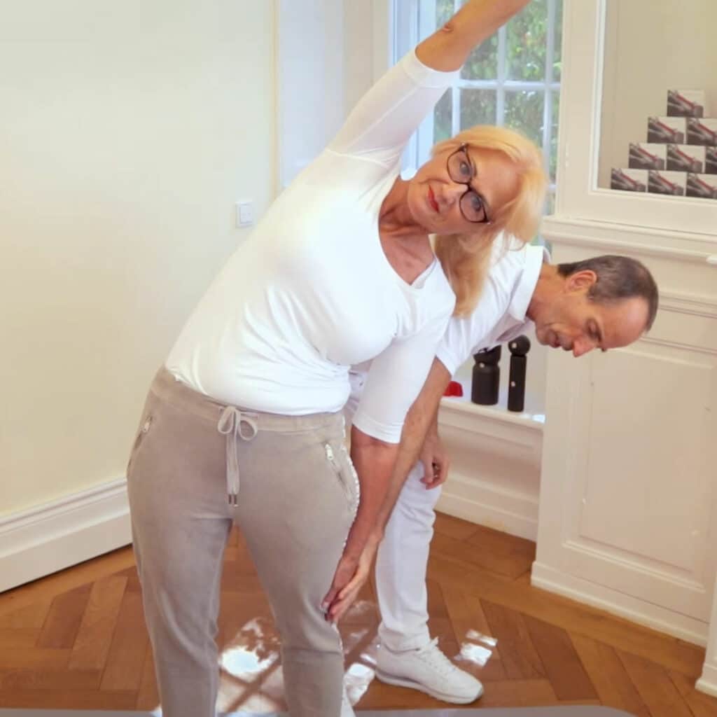 Eine Frau in Trainingskleidung führt die Übung nach Anweisung von Schmerzspezialist Roland Liebscher-Bracht aus. Sie lehnt sich zur Seite nach unten, ein Arm ist lang über ihren Kopf gestreckt und der andere liegt an ihrem Oberschenkel. Roland Liebscher-Bracht steht hinter ihr und korrigiert ihre Position.