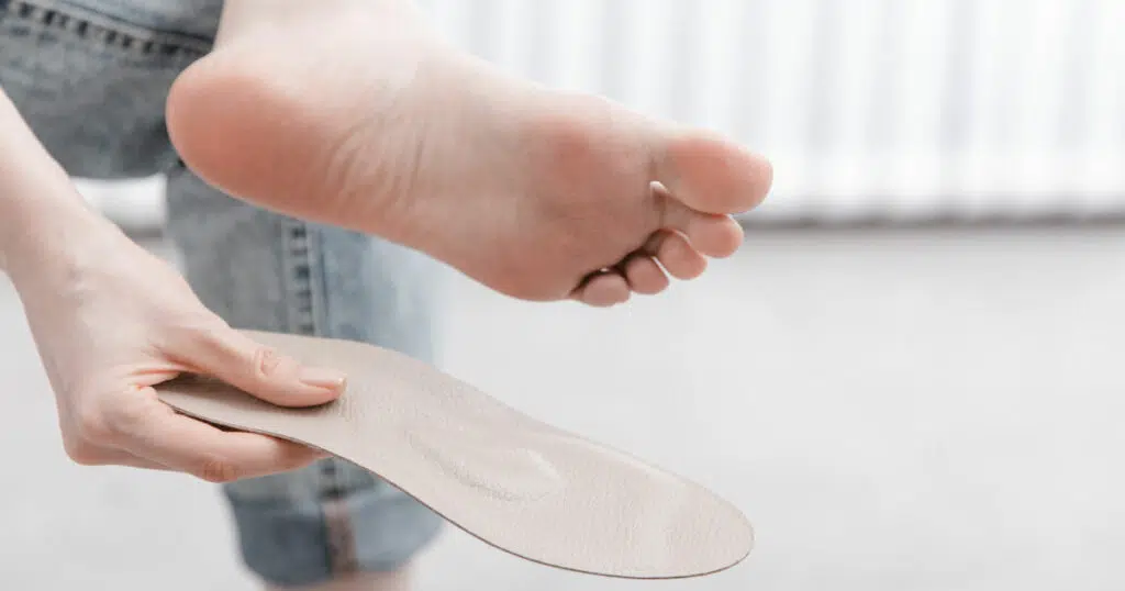 Eine Frau hat ihr linkes Bein im Sitzen über das rechte gelegt. In ihrer rechten Hand hält sie eine Einlegesohle, während ihre linke Fußsohle zu sehen ist.