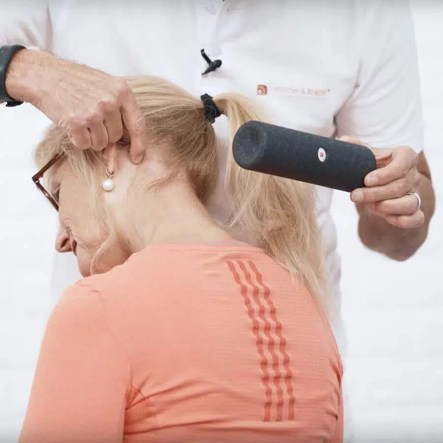 Eine Person sitzt und neigt den Kopf nach vorne unten rechts. Eine stehende Person in weißer Kleidung hat eine Faszienrolle in der Hand und zeigt auf einen Punkt am Kopf der sitzenden Person.